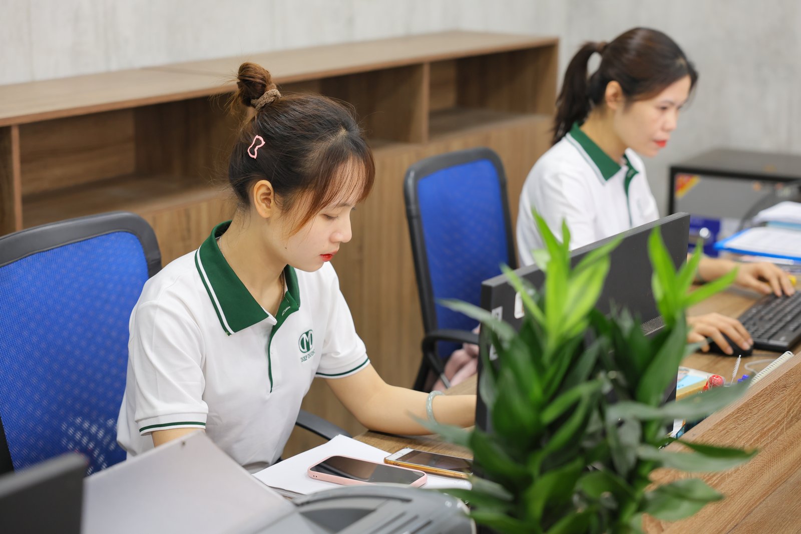 Hai nữ nhân viên Điệp Dương mặc áo đồng phục trắng cổ xanh, làm việc bên máy tính tại bàn gỗ